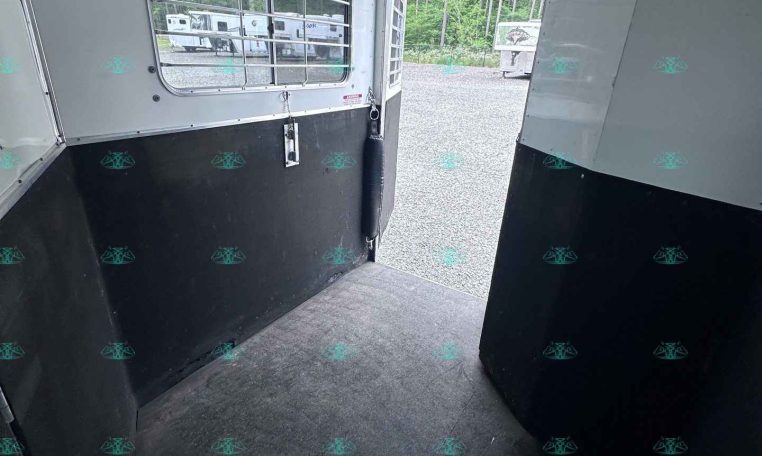 Interior of a horse trailer with white walls and black lower panels, window with bars, and open rear door to a gravel lot with trees and other trailers outside.