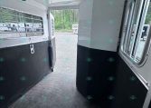 Interior of a horse trailer with a divider panel and windowed side walls, open rear door to a gravel lot.