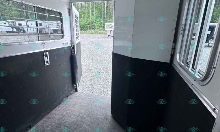Interior of a horse trailer with a divider panel and windowed side walls, open rear door to a gravel lot.