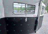 Interior of a horse trailer with a barred window, white walls, and a black lower panel; other trailers are visible outside.