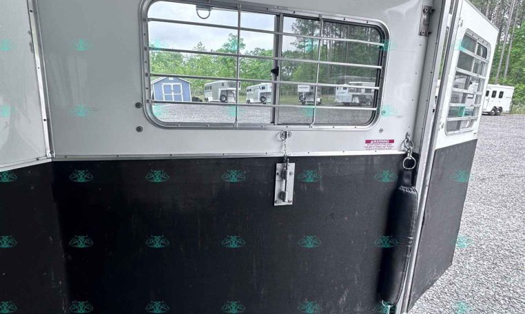 Interior of a horse trailer with a barred window, white walls, and a black lower panel; other trailers are visible outside.