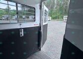 Interior of a plain horse trailer with a metal window grille and open door, view to a gravel lot and trees beyond.