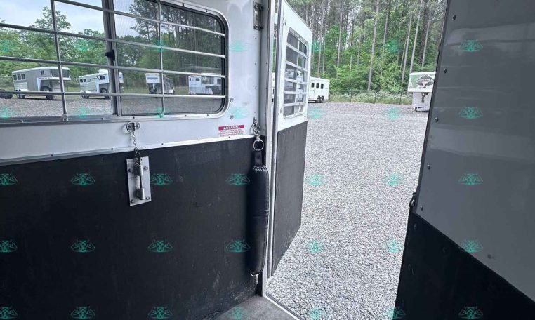 Interior of a plain horse trailer with a metal window grille and open door, view to a gravel lot and trees beyond.