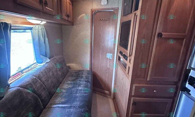 Interior of a camper trailer: wooden cabinets, a blue-patterned sofa, window with blue curtains, and an overhead air vent/ventillator.