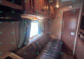 Wood-panelled RV living area with a brown sofa, overhead cabinets, and a window with blue curtains