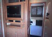RV kitchenette with wooden cabinets, microwave above an oven, and a compact fridge to the right near the entry door with a window shade.