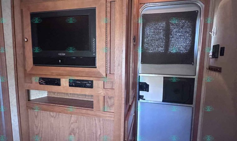 RV kitchenette with wooden cabinets, microwave above an oven, and a compact fridge to the right near the entry door with a window shade.