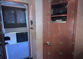 Trailer interior showing a wooden cabinet with a built-in microwave and a tall refrigerator beneath; a doorway to the side and a fire extinguisher on the floor.
