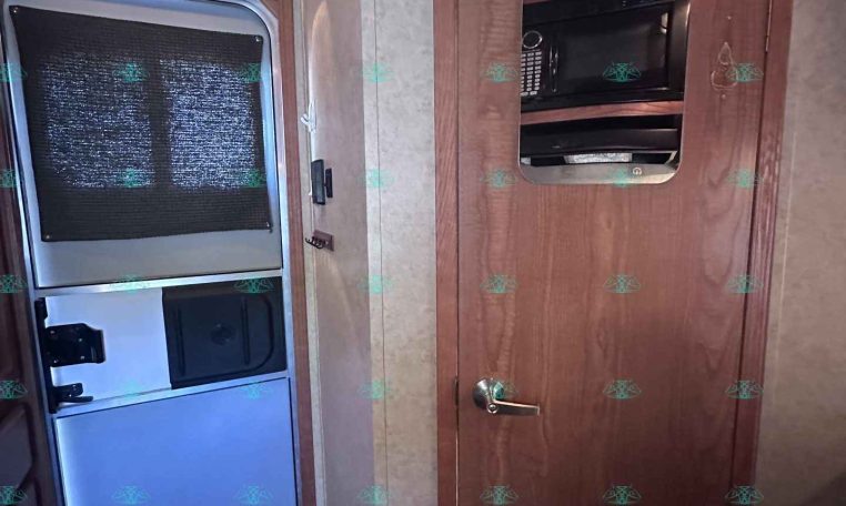 Trailer interior showing a wooden cabinet with a built-in microwave and a tall refrigerator beneath; a doorway to the side and a fire extinguisher on the floor.