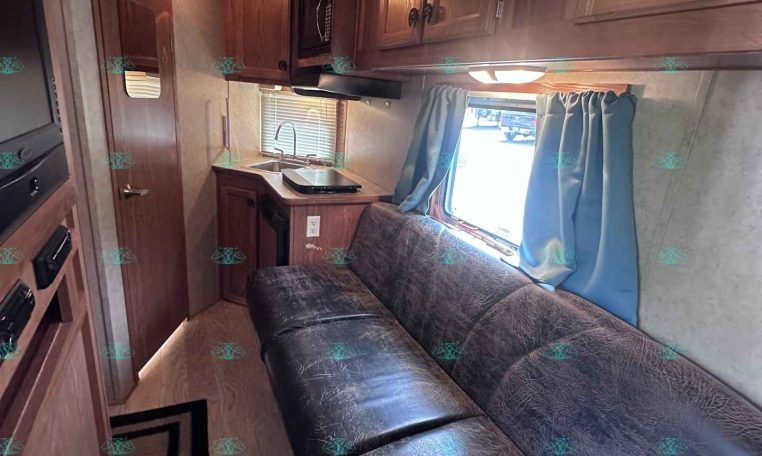 Interior of a compact RV/kitchen area with wooden cabinets, microwave, sink, stove, and a sofa under a window with blue curtains far end