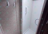 Compact RV shower stall with white interior, built-in curved grab bar, and a skylight above the door opening from the bathroom.