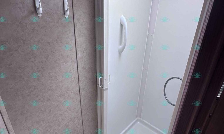 Compact RV shower stall with white interior, built-in curved grab bar, and a skylight above the door opening from the bathroom.