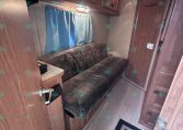 Interior of a small RV living area with brown wood cabinets above a brown upholstered sofa and a light blue curtain along the window