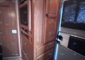 Wooden RV kitchen area with tall pantry cabinets and a built-in microwave, seen beside a window with blinds to the right.