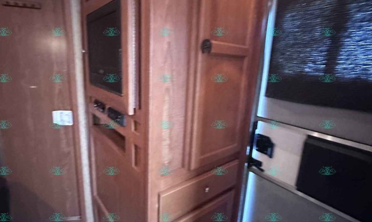 Wooden RV kitchen area with tall pantry cabinets and a built-in microwave, seen beside a window with blinds to the right.