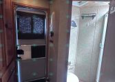 Interior of an RV kitchen with a tall stainless steel refrigerator beside wooden cabinets, doorway to a bathroom visible with a glass shower door and toilet.