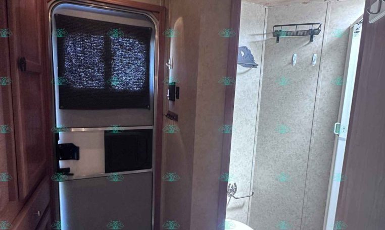 Interior of an RV kitchen with a tall stainless steel refrigerator beside wooden cabinets, doorway to a bathroom visible with a glass shower door and toilet.