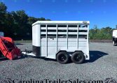 White livestock trailer on a gravel lot with trees and a red tractor attachment nearby under a blue sky, a dealership setting.