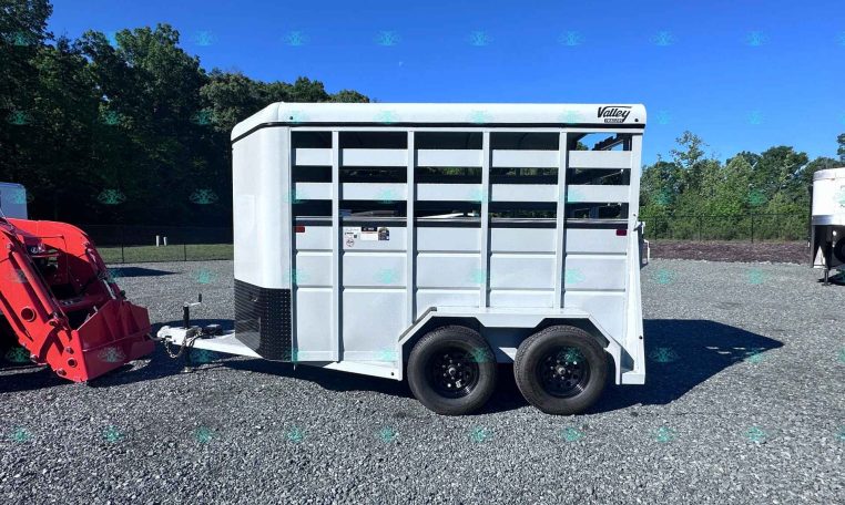 White livestock trailer on a gravel lot with trees and a red tractor attachment nearby under a blue sky, a dealership setting.
