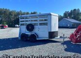 White livestock trailer with a spare tire mounted on the side sits on a gravel lot, trees in the background and a blue building nearby.