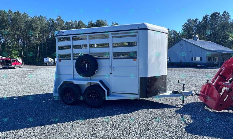 White livestock trailer with a spare tire mounted on the side sits on a gravel lot, trees in the background and a blue building nearby.