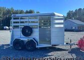 White livestock trailer with a mounted spare tire on the side, parked on a gravel lot with trees and a blue sky in the background.