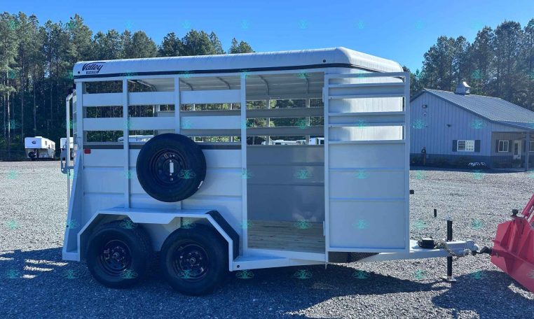 White livestock trailer with a mounted spare tire on the side, parked on a gravel lot with trees and a blue sky in the background.