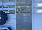 White aluminum trailer with open slatted side, showing interior stalls and a spare tire mounted on the outside. Clear blue sky above.