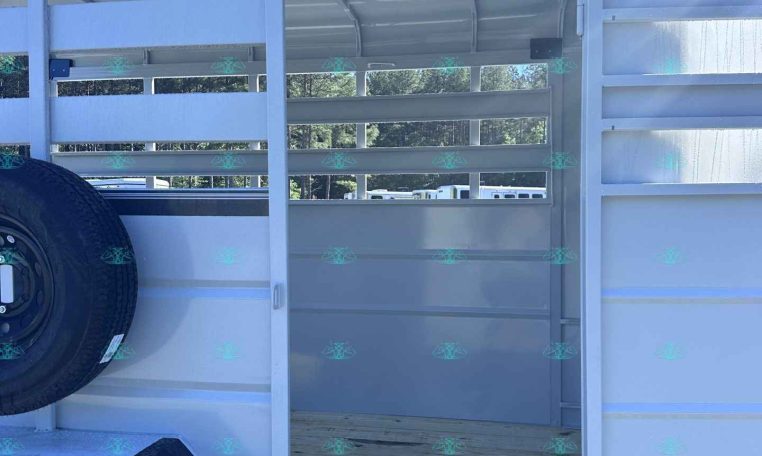 White aluminum trailer with open slatted side, showing interior stalls and a spare tire mounted on the outside. Clear blue sky above.