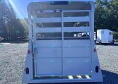 Rear view of a white livestock trailer with its ramp down, parked on a gravel lot under a clear blue sky with trees in the background.