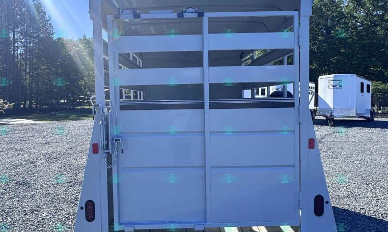 Rear view of a white livestock trailer with its ramp down, parked on a gravel lot under a clear blue sky with trees in the background.