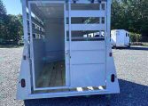 Open-backed white livestock trailer with wooden floor and interior gates, parked on gravel with another trailer in the background.