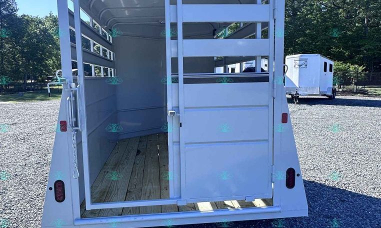 Open-backed white livestock trailer with wooden floor and interior gates, parked on gravel with another trailer in the background.