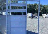 White metal trailer ramp and gate in a gravel lot, with other horse trailers and trees in the background on a sunny day.