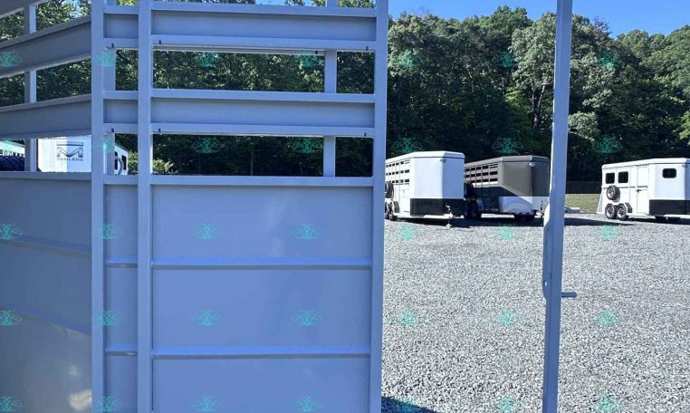 White metal trailer ramp and gate in a gravel lot, with other horse trailers and trees in the background on a sunny day.