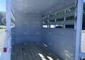 Interior of an empty horse trailer with wooden floor and white metal walls, rear doors open for loading.
