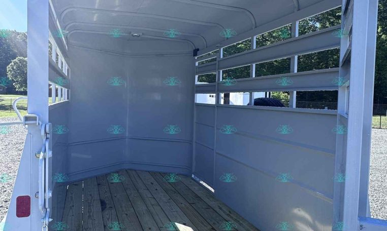 Interior of an empty horse trailer with wooden floor and white metal walls, rear doors open for loading.