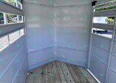 Interior of a white horse trailer with a wooden floor and open side doors, showing horizontal slatted windows and light from outside