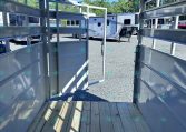 Interior of a metal trailer with an open rear ramp, looking out at a gravel lot with parked travel trailers.