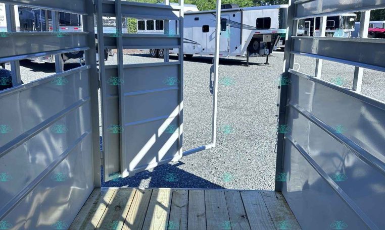 Interior of a metal trailer with an open rear ramp, looking out at a gravel lot with parked travel trailers.