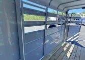 Interior of a gray metal horse trailer with partition rails and a wooden plank floor, sunlight visible through open side gate.