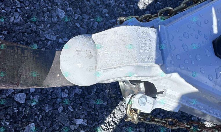 Close-up of a trailer hitch coupler with safety chains on a rocky gravel surface.