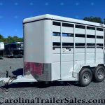 Silver livestock trailer with side slats and dual wheels, parked on a gravel lot with other trailers and trees in the background