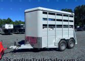Silver livestock trailer with side slats and dual wheels, parked on a gravel lot with other trailers and trees in the background