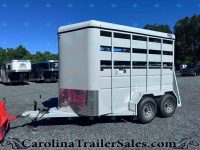 Silver livestock trailer with side slats and dual wheels, parked on a gravel lot with other trailers and trees in the background