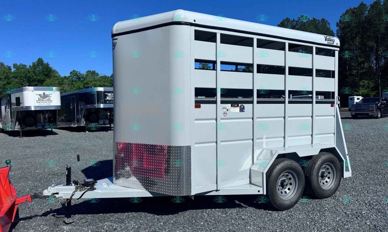 Silver livestock trailer with side slats and dual wheels, parked on a gravel lot with other trailers and trees in the background