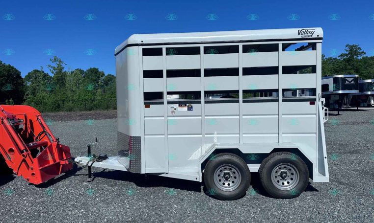 White Valley Trailers livestock trailer parked on a gravel lot with a red equipment attachment in the foreground and trees in the background
