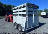 White livestock trailer with side vents and dual rear doors hitched to a red tractor on a gravel lot.