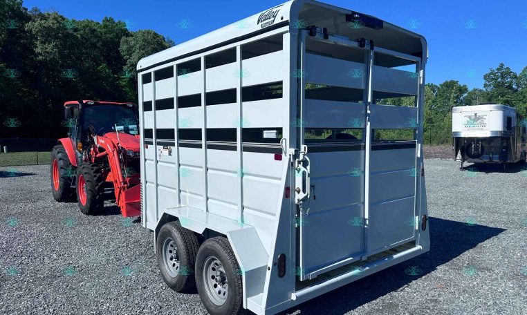 White livestock trailer with side vents and dual rear doors hitched to a red tractor on a gravel lot.