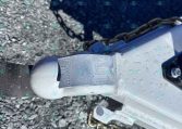 Close-up of a trailer coupler and safety chains on a gravel surface, showing the hitch latch and part of the trailer frame.
